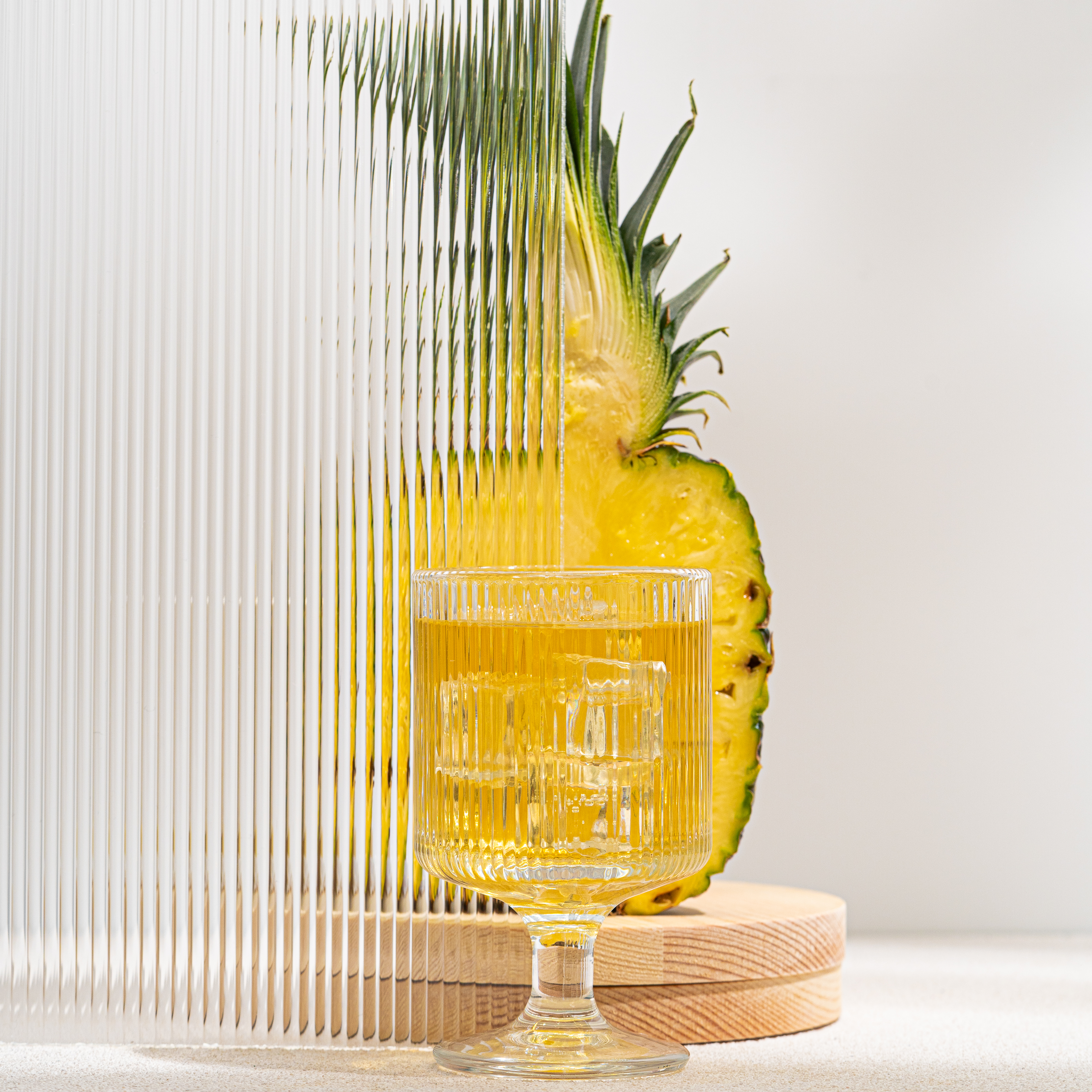 Halved pineapple, where half is blocked by a texted glass; glass cup of tea with ice cubes sitting in front of the pineapple