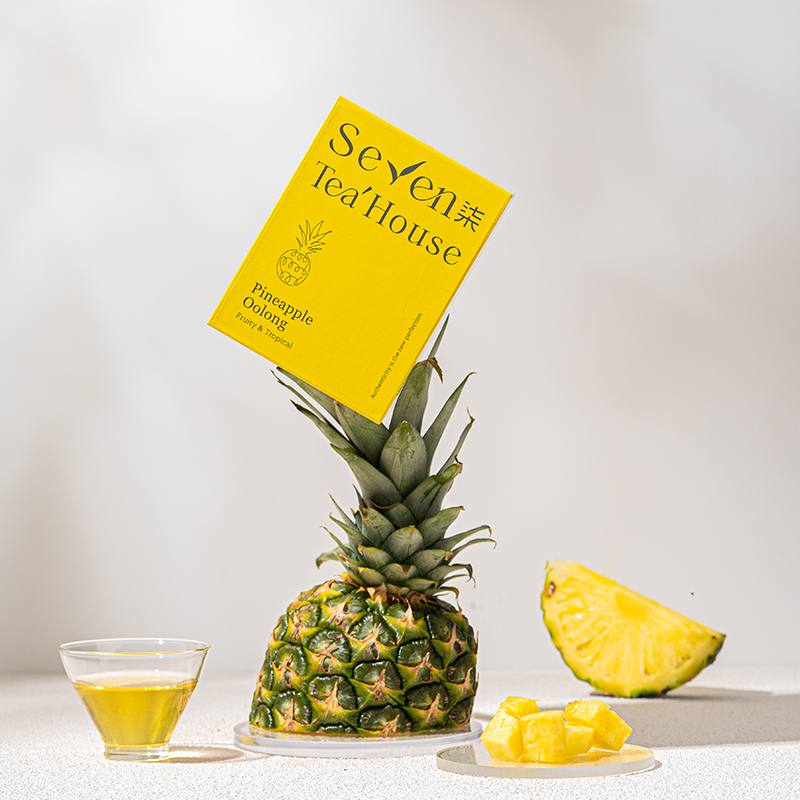 A yellow box of pineapple oolong tea resting on top of a pineapple with cut pineapple and a glass of tea on the side