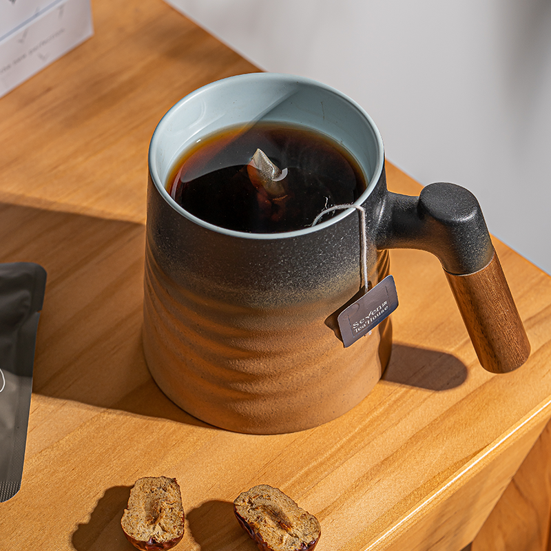 A clay mug with brewed tea and a tea bag sitting on a wooden surface surrounded by tea accessories