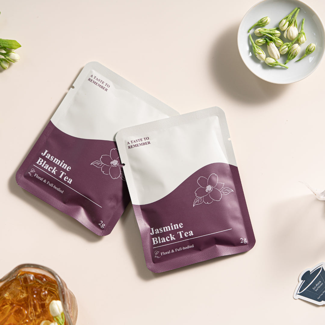 Two packages of Jasmine Black Tea on a light beige background with a plate of jasmine buds on the top right and cup of tea on the bottom left